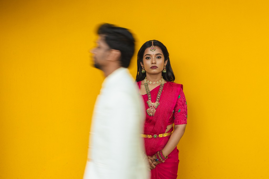 Pre-Wedding — Bride in red saree against bold yellow wall, groom blurred in foreground